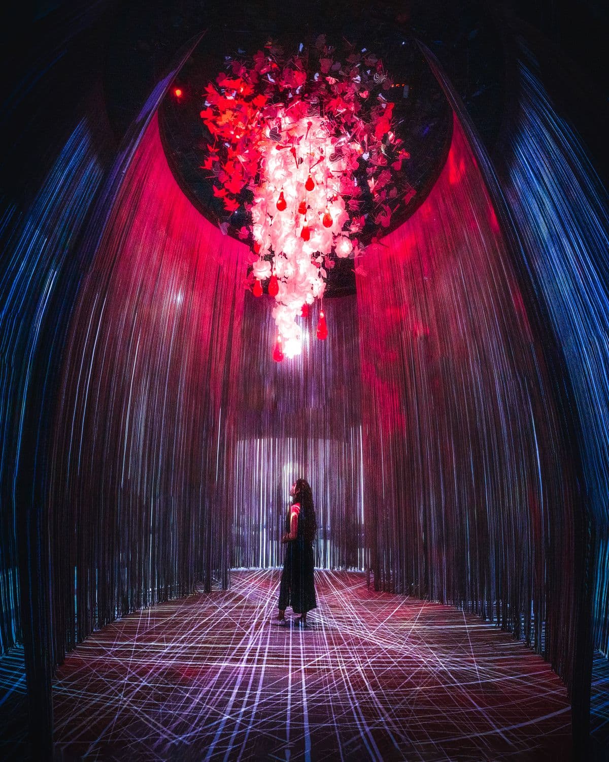A visitor stands beneath a red and blue chandelier installation in a dark exhibition room.