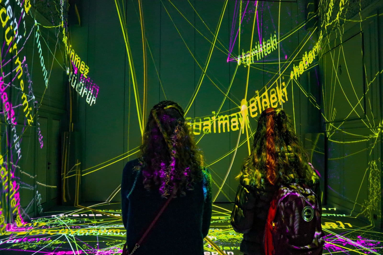Two visitors cross a dark gallery filled with green, yellow, and magenta projected text.