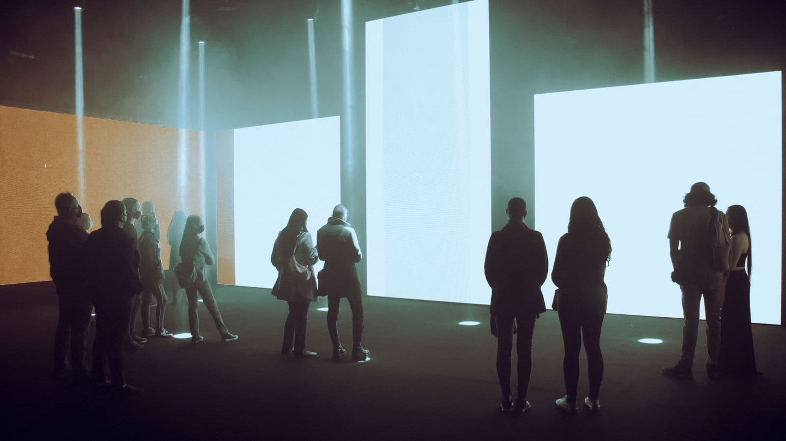 Visitors appear in silhouette before a row of luminous screens in a museum gallery.
